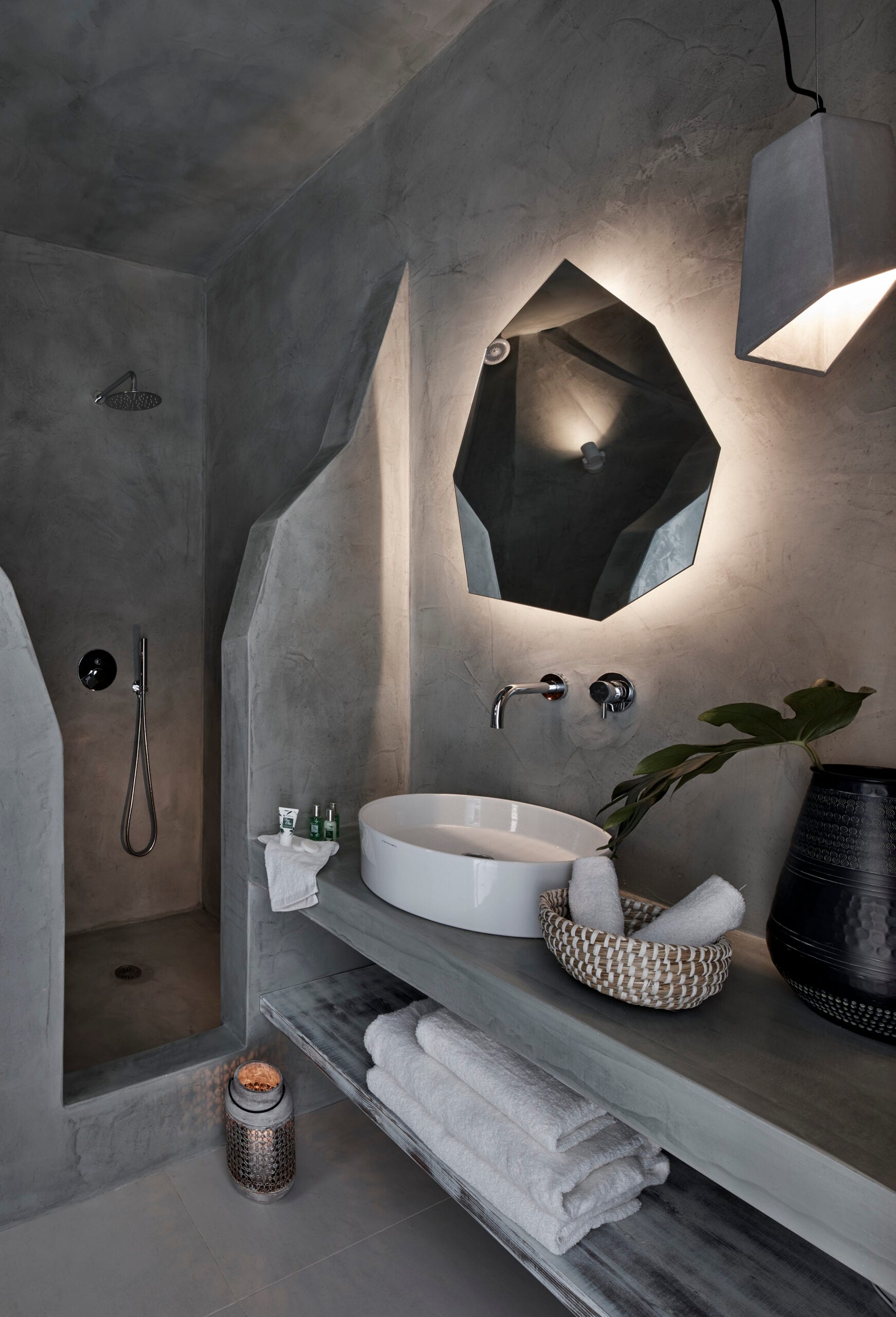 Sculpted grey microcement bathroom with a backlit faceted mirror and rainfall shower