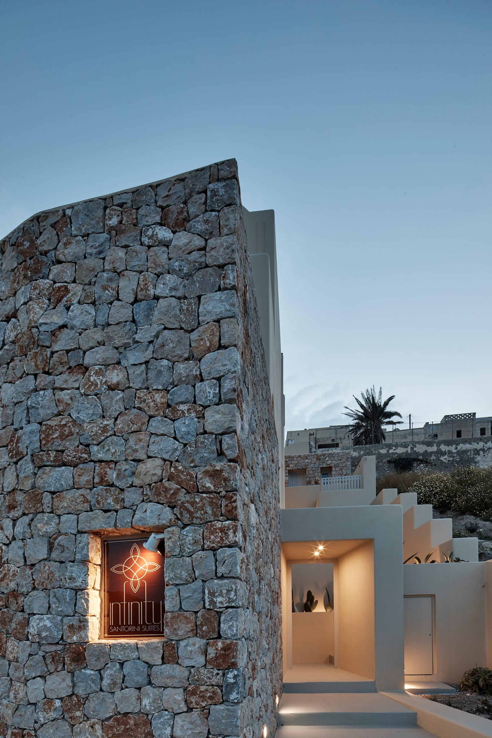 Suite One entrance at dusk, a rugged natural-stone tower beside the whitewashed Cycladic walkway