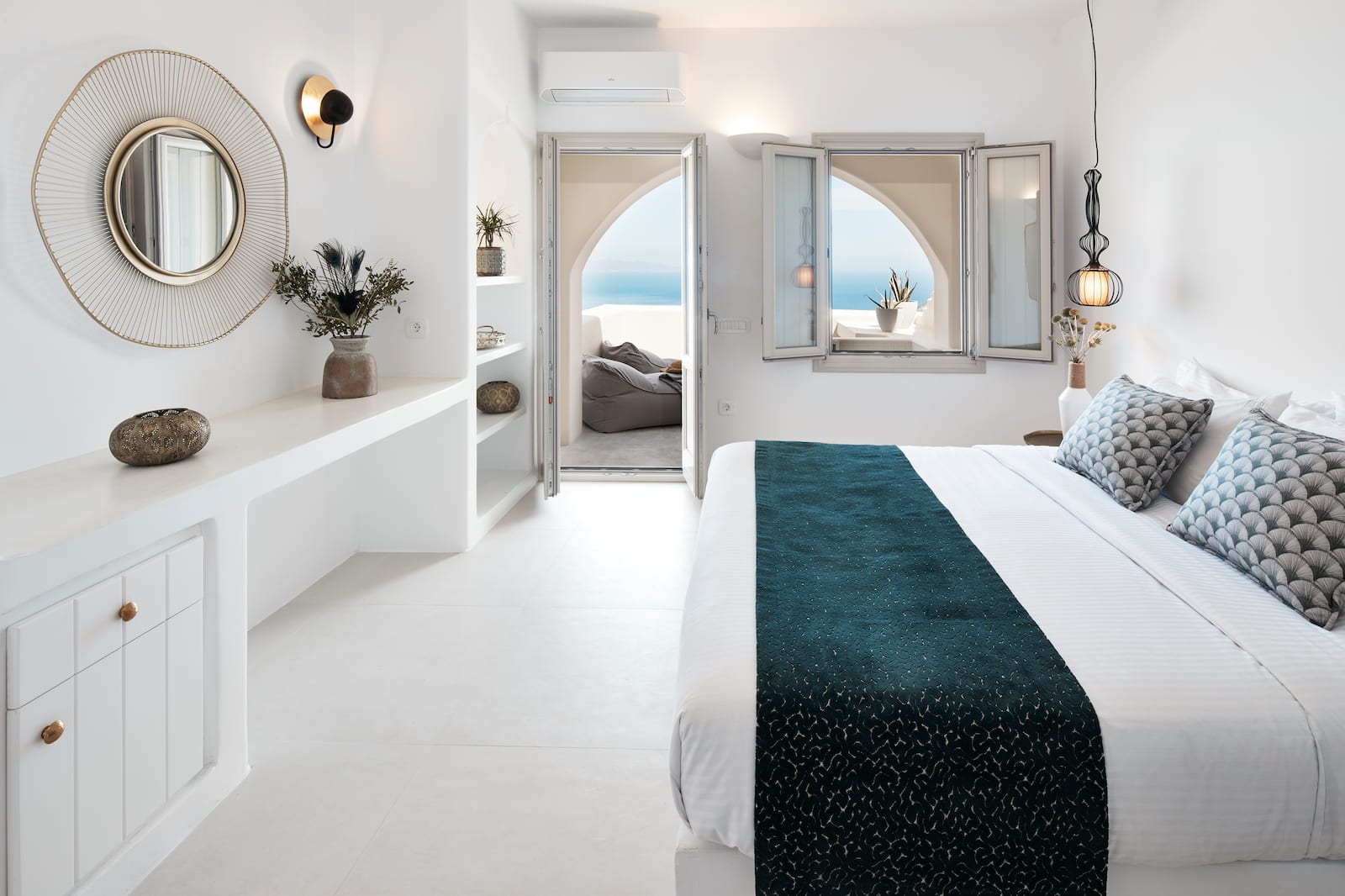 Bedroom with a round gold mirror, whitewashed vanity and an arched doorway framing the Aegean Sea