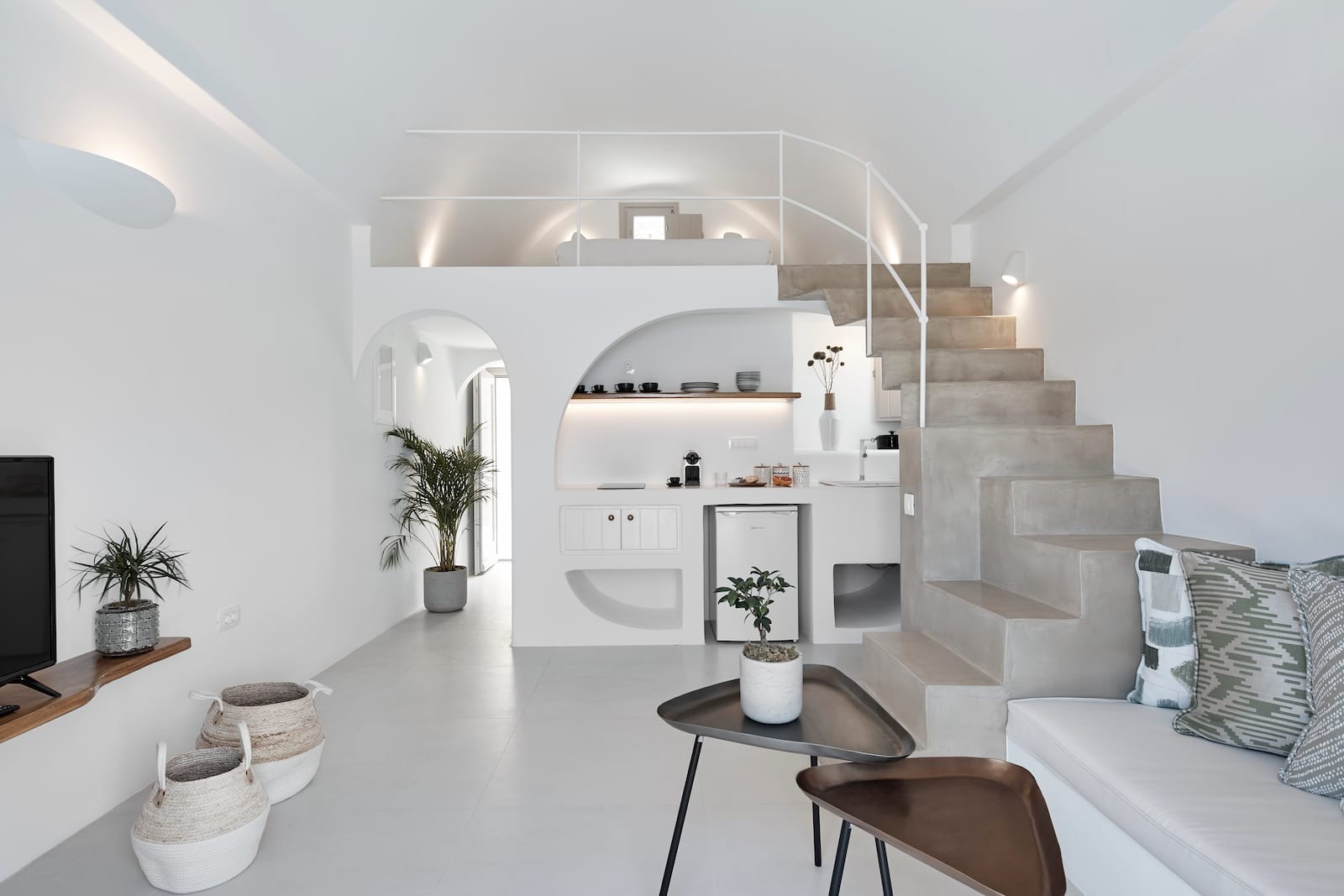 Bright open-plan living area with a sculptural concrete staircase, arched kitchenette niche and loft mezzanine above
