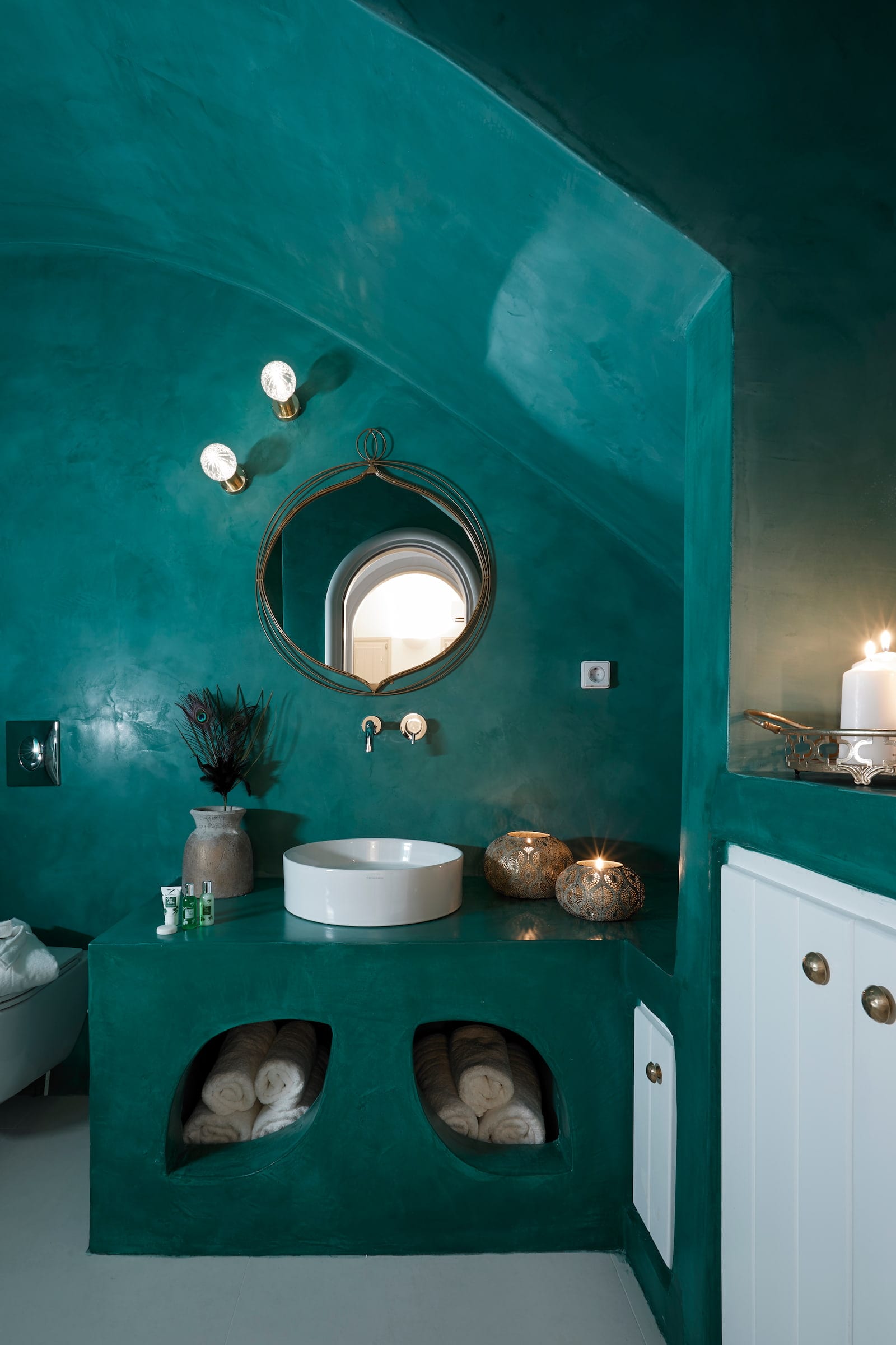 Emerald plaster bathroom with a round brass mirror, white vessel basin and towels tucked into curved niches