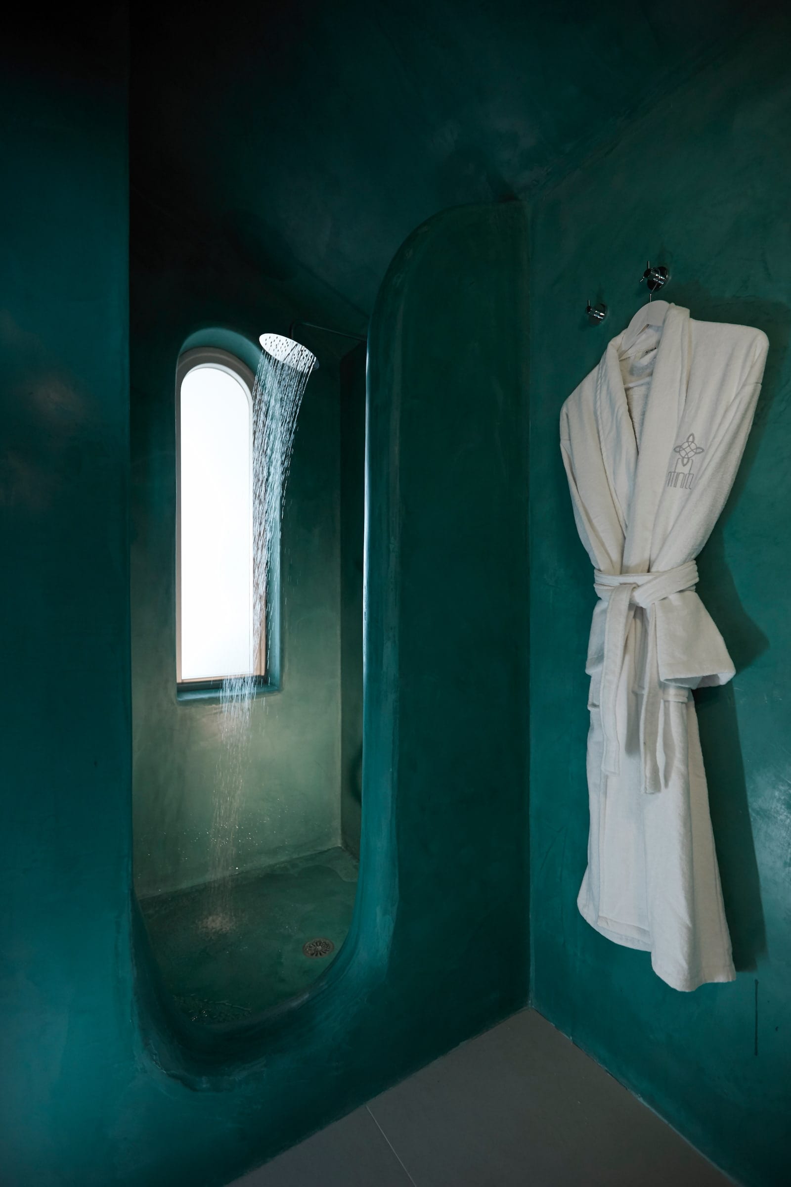 Teal tadelakt walk-in shower with a rainfall head beside an arched window and hanging robe