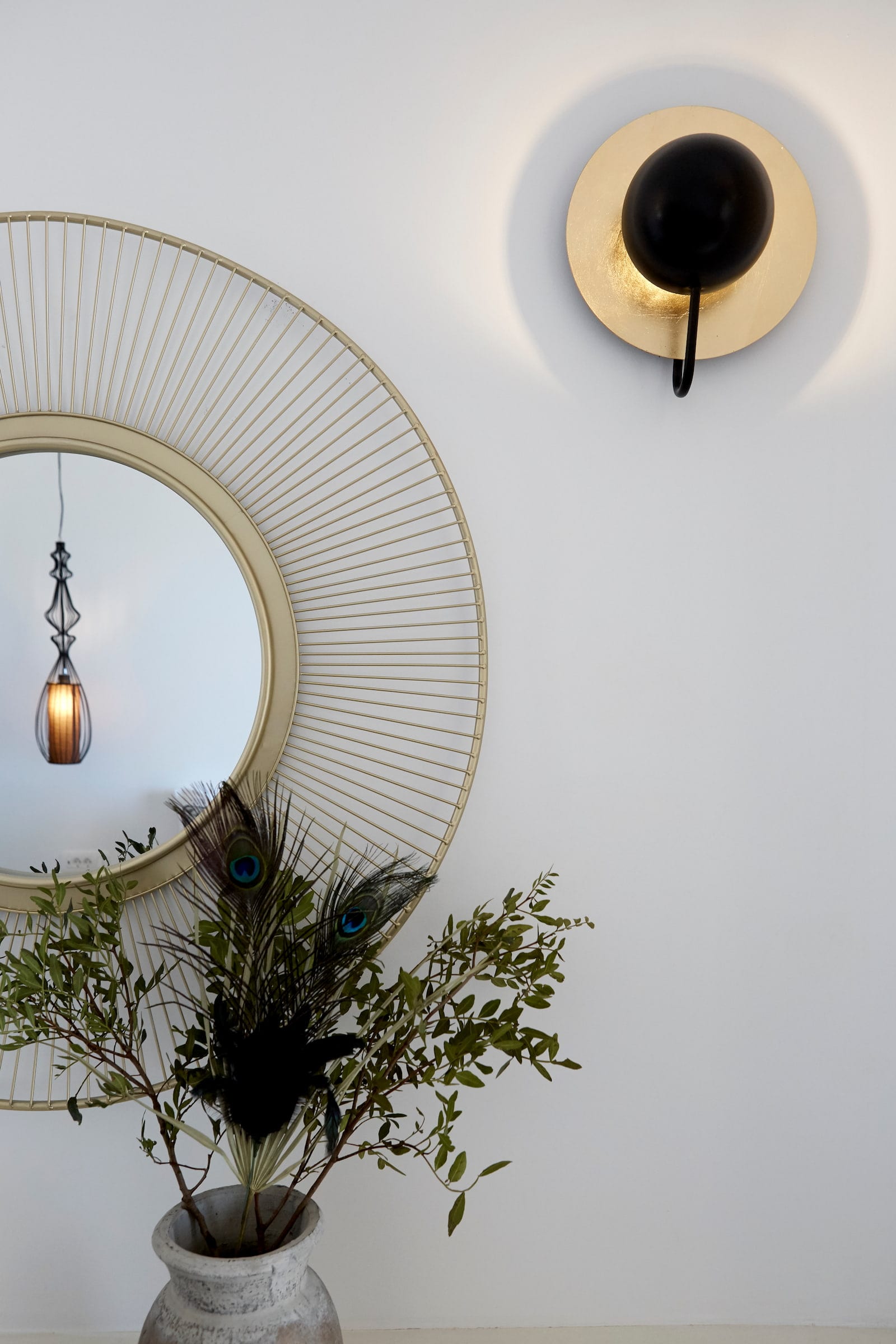 Gold wire sunburst mirror and a black-and-brass wall sconce above a vase of peacock feathers