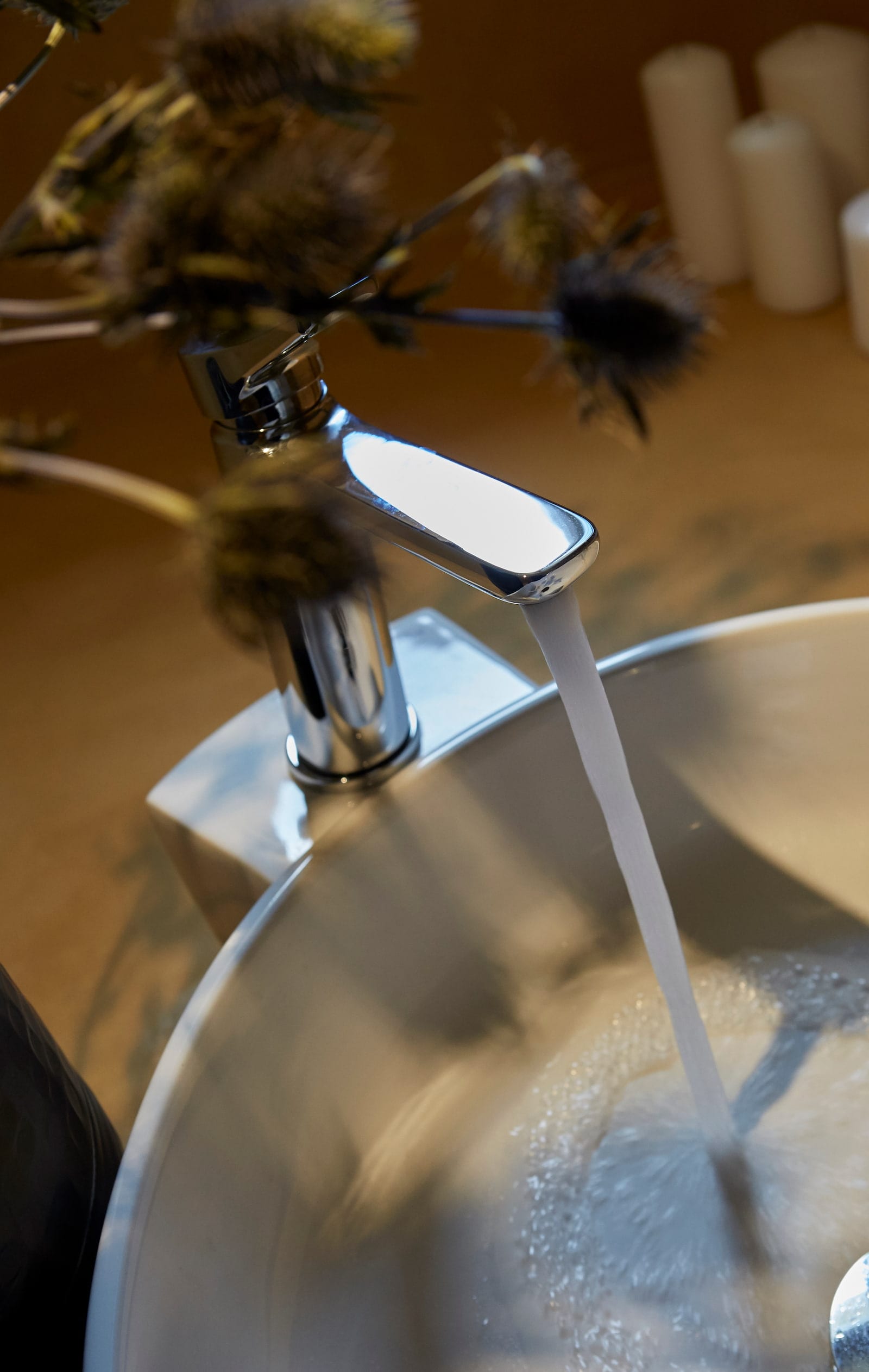 Polished chrome tap running water into a white basin beside dried thistle stems