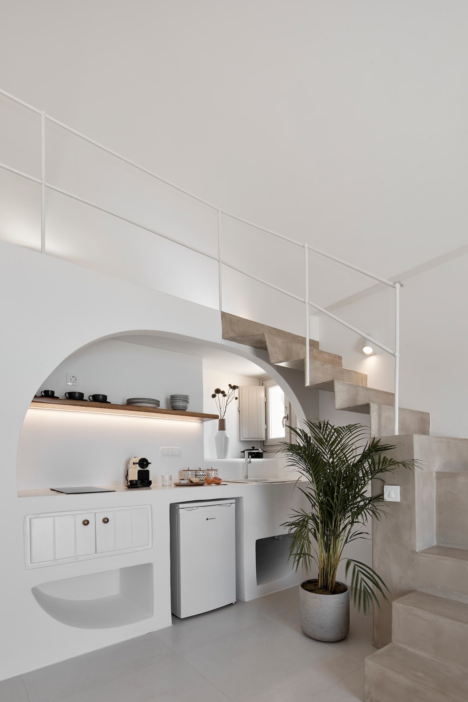 Arched kitchenette with a mini-fridge and open shelf beneath the floating concrete stairs and loft
