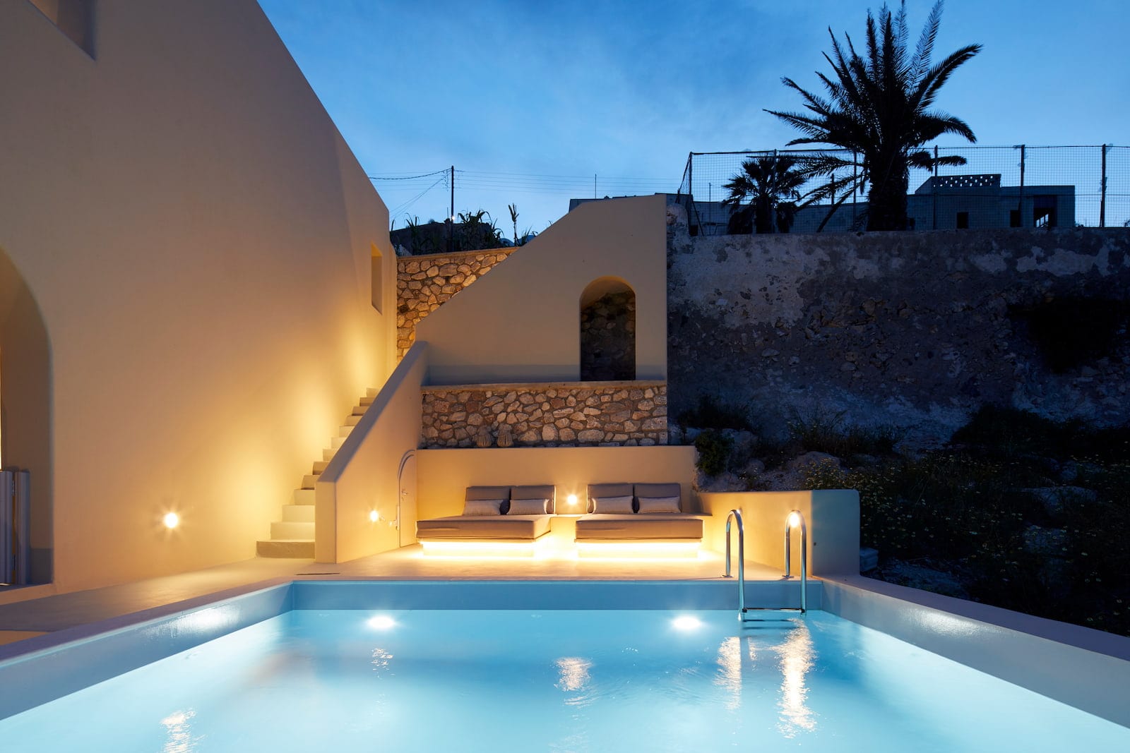 Illuminated pool at twilight with cushioned sunbeds set against traditional Cycladic stone terracing