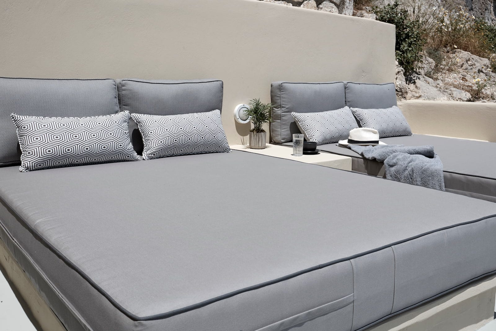 Outdoor terrace daybed with grey cushions, geometric pillows and a sun hat beside rugged rock