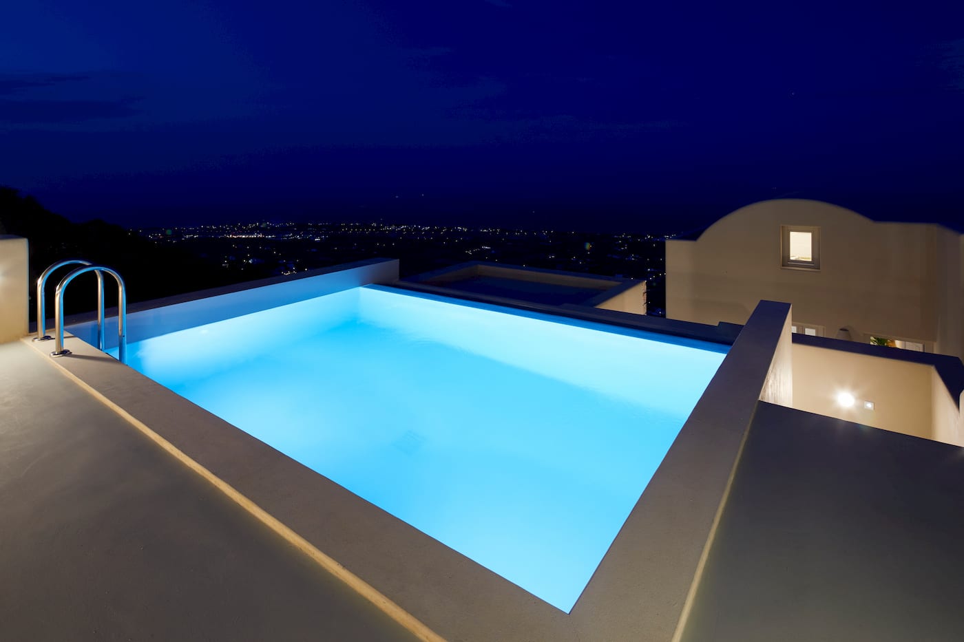 Illuminated pool glowing turquoise at night above the twinkling lights of Santorini below