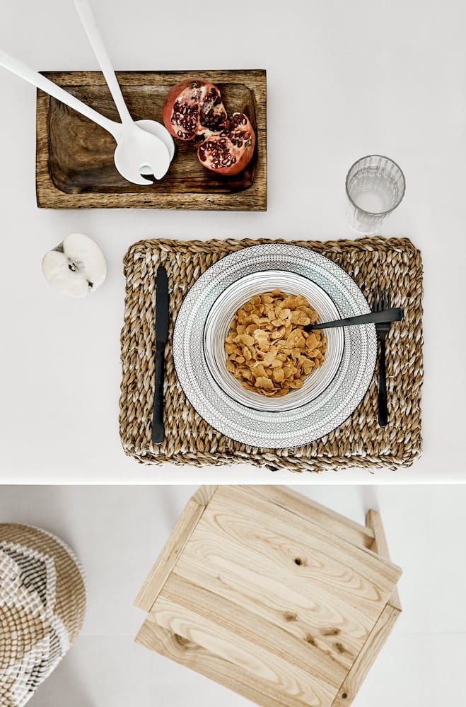 Overhead breakfast flat-lay with a cereal bowl, woven placemat and pomegranate on a wooden table