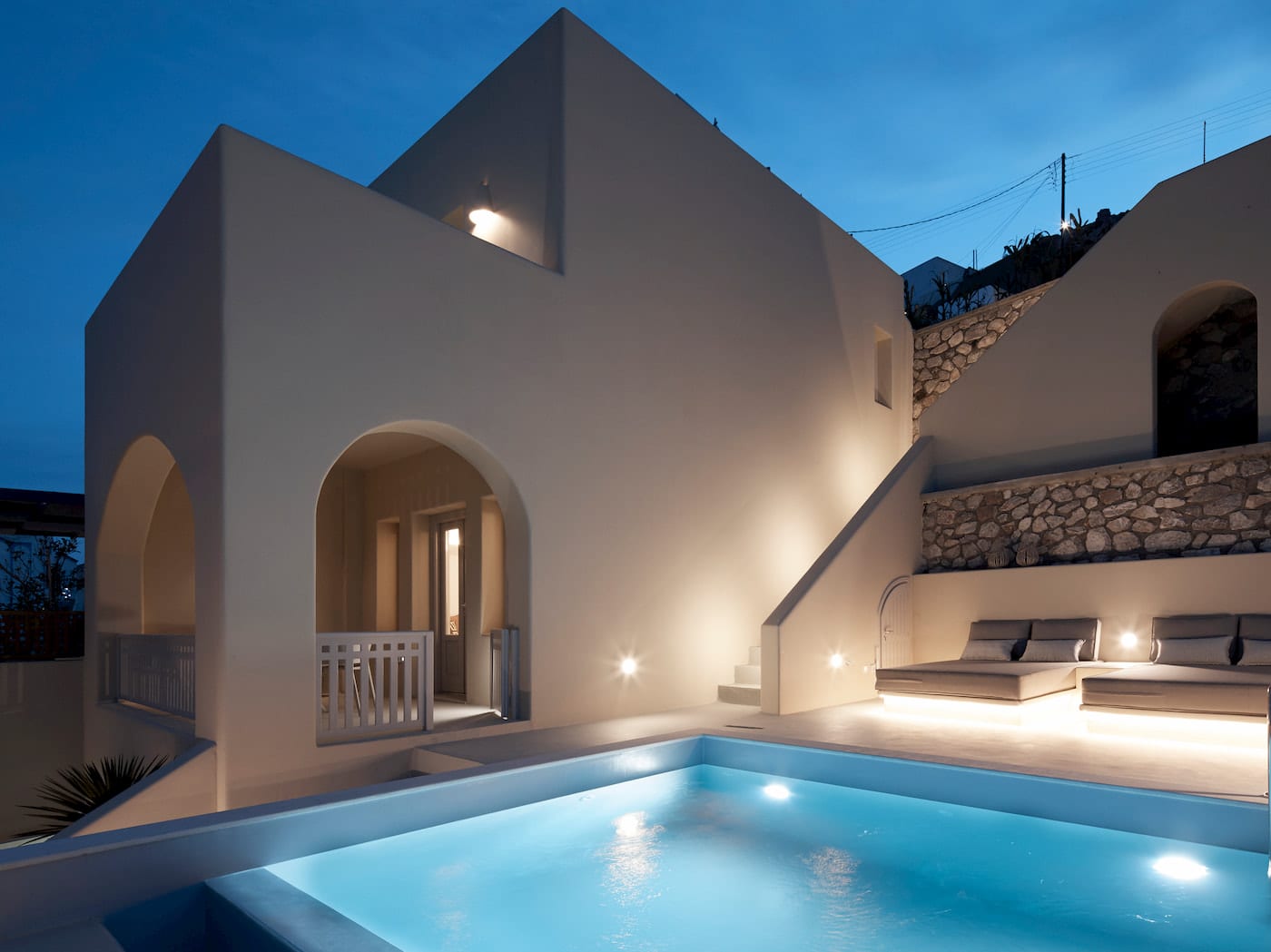 Whitewashed Cycladic exterior at dusk with a lit plunge pool, stone wall and cushioned loungers