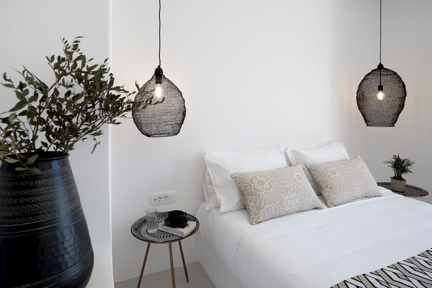 Minimal bedroom with white linens, twin black wire pendants and an olive-branch arrangement