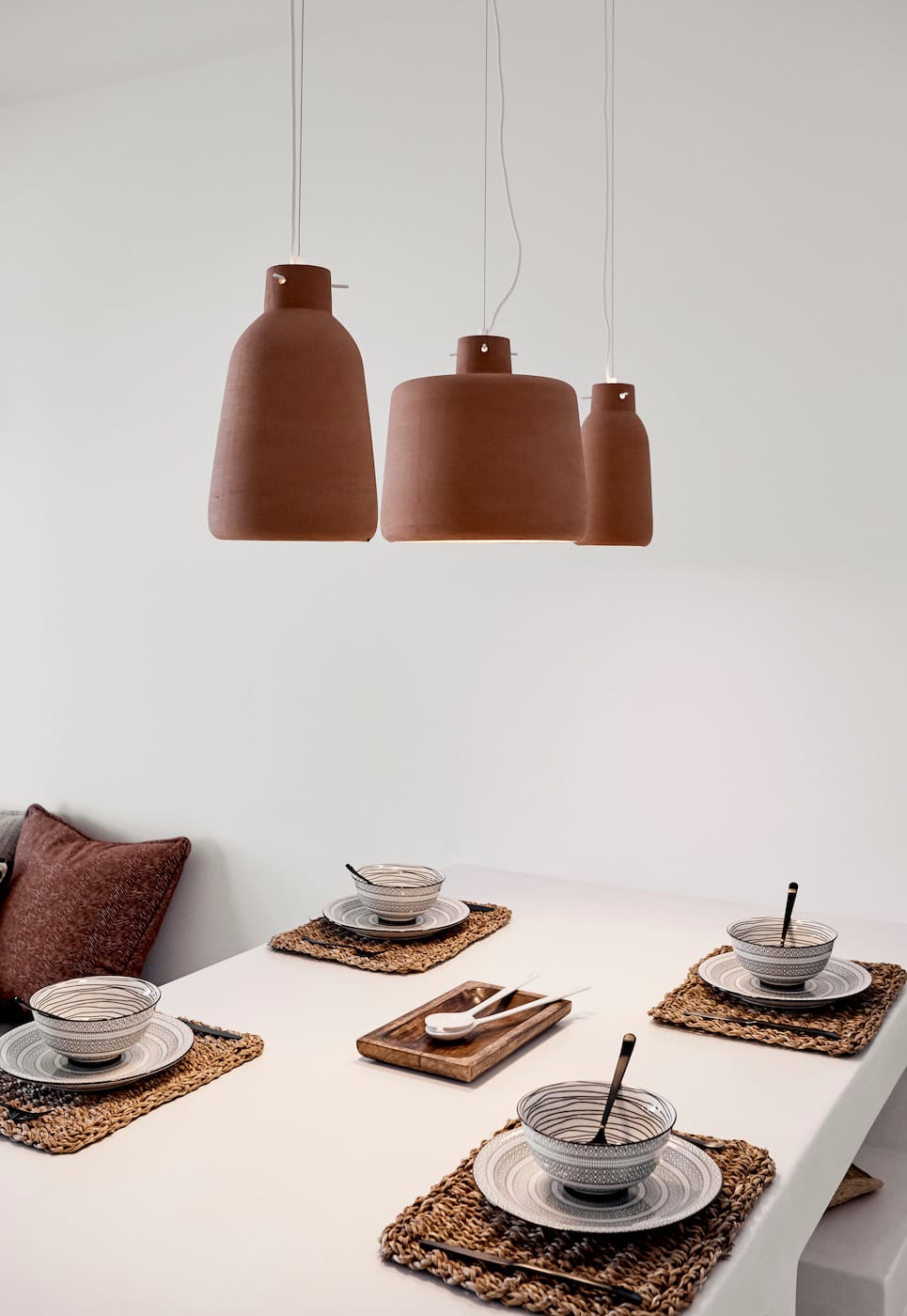 Dining table dressed with patterned bowls and woven placemats beneath three terracotta pendant lamps